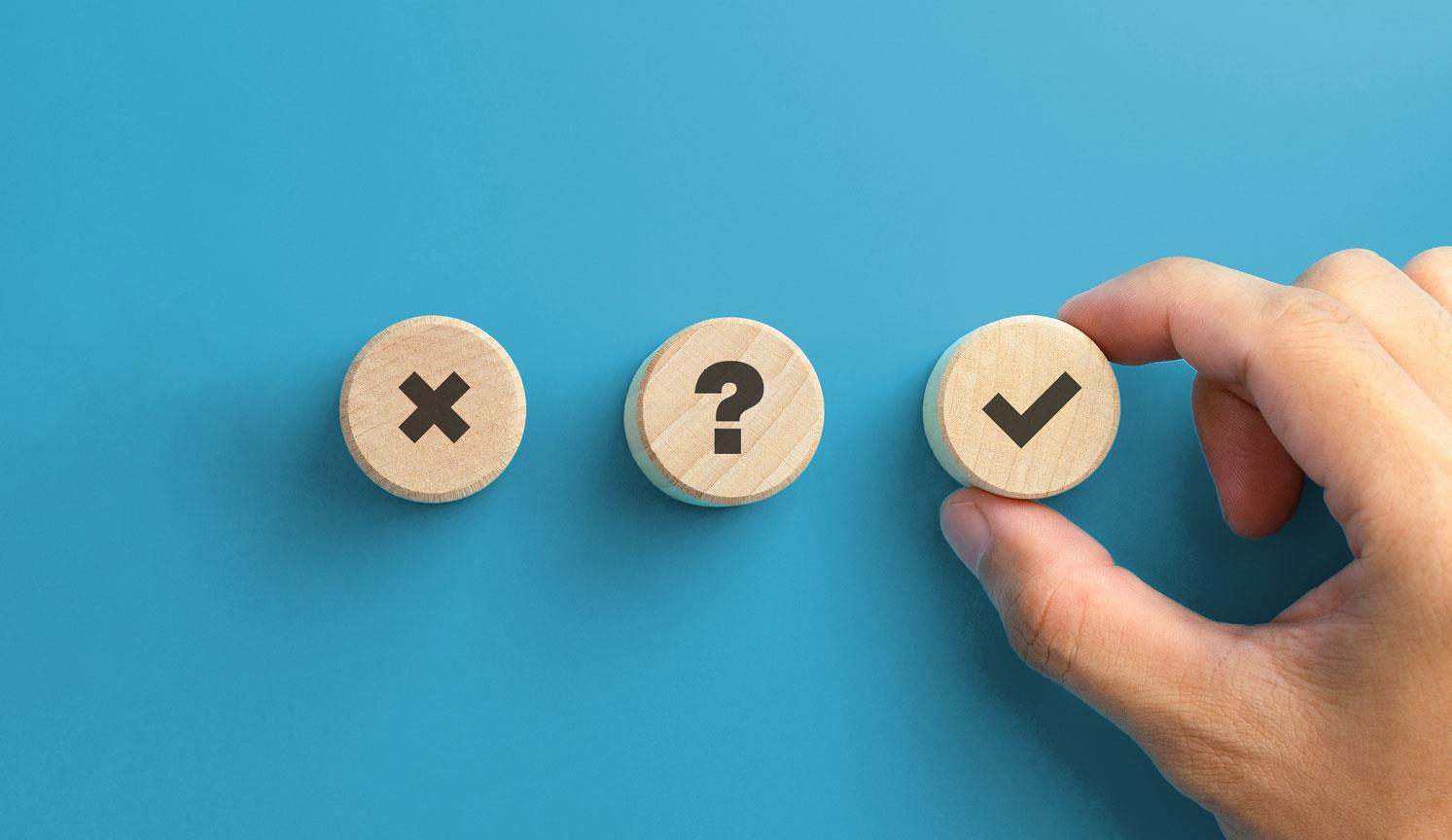 3 wooden counters, with a cross, a tick and a question mark, fingers grasping the one with the tick